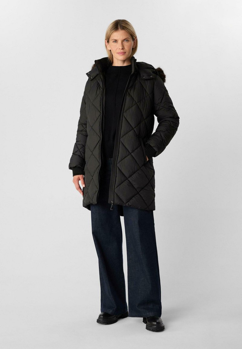 Cappotto imbottito nero a trapunte con cappuccio e bordo in pelliccia sintetica, dotato di zip frontale e tasche laterali, indossato sopra un maglione nero e jeans scuri.