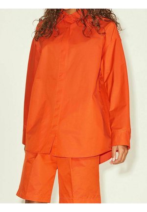 Kvinde iført løstsiddende, knaldrød langærmet skjorte og matchende orange shorts til knæene, står mod en ensfarvet baggrund.