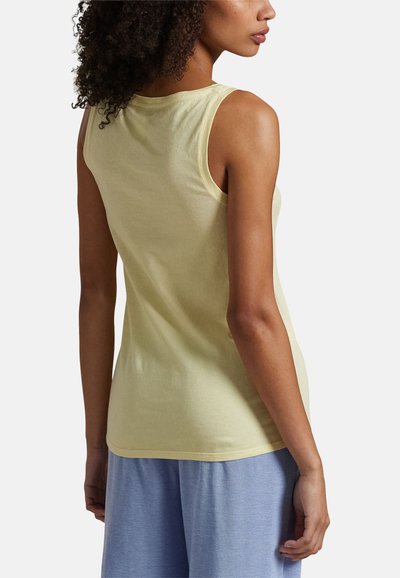 Femme aux cheveux bouclés portant un haut sans manches jaune clair et un pantalon ample bleu clair, vue de dos sur un fond uni.