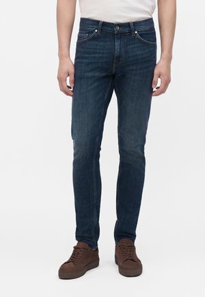 Mand iført slim-fit mørkeblå jeans og brune sneakers, stående mod en ensfarvet lys baggrund.