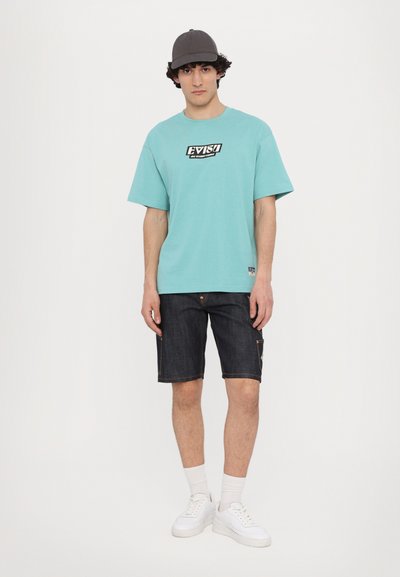 Τυρκουάζ oversized T-shirt με λογότυπο μπροστά, σε συνδυασμό με σκούρες τζιν βερμούδες. Το μοντέλο φοράει λευκά αθλητικά και γκρι καπέλο. Απλός και καθημερινός σχεδιασμός.