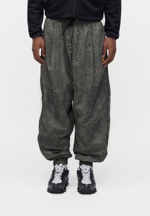 Mies, jolla on harmaat leveät housut naruilla vyötäröllä, mustavalkoiset Nike Shox -tennarit, seisoo yksinkertaisen valkoisen taustan edessä.