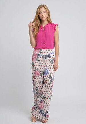 Mujer joven con cabello largo y rubio que lleva una blusa sin mangas de color rosa brillante y pantalones anchos blancos con lunares rosas y estampado floral.
