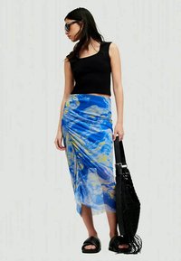 Μπλε και κίτρινη φούστα περιτυλίγματος με μοτίβο, διαφανές ύφασμα, σχεδίαση με κρέμασμα, συνδυασμένη με ένα μαύρο crop top, μαύρες παντόφλες και μια μαύρη τσάντα tote.