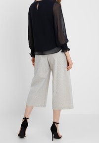 Blusa nera trasparente a maniche lunghe con apertura a chiave sulla schiena, abbinata a culotte bianche a righe e sandali con tacco neri.