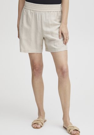 Beige, gerippte Shorts mit einem elastischen Bund, die einen lockeren Schnitt und eine glatte Textur aufweisen, kombiniert mit neutralen Rutsch-Sandalen.