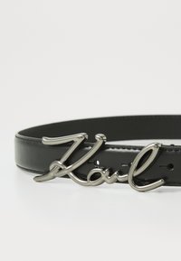 Zwarte leren riem met een glanzende metalen gesp waarop in cursieve letters "Karl" staat. Inclusief verschillende verstelgaten langs de lengte.