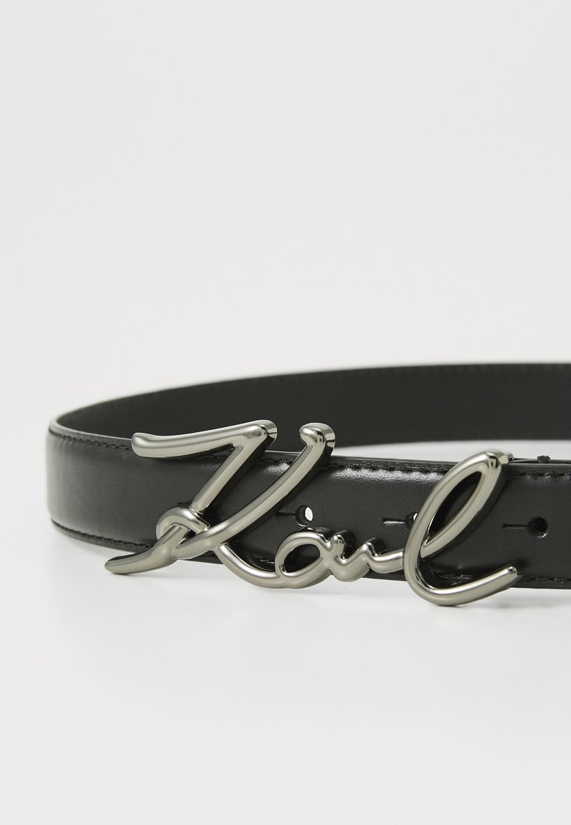 Ceinture en cuir noir avec une boucle en métal brillante portant le texte en cursive "Karl". Elle comprend plusieurs trous d'ajustement sur toute sa longueur.
