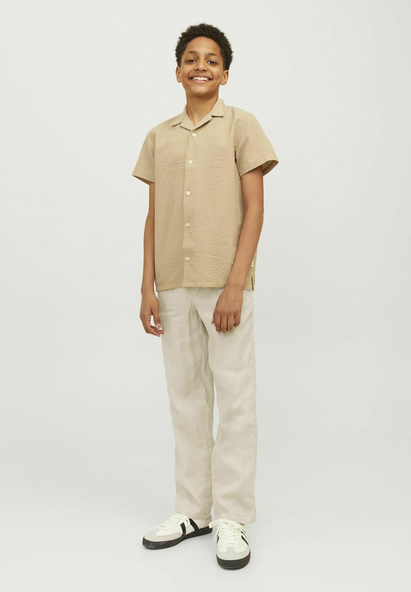 Beige overhemd met korte mouwen en een gestructureerd patroon, gecombineerd met lichtgekleurde broeken en witte sneakers met zwarte accenten.