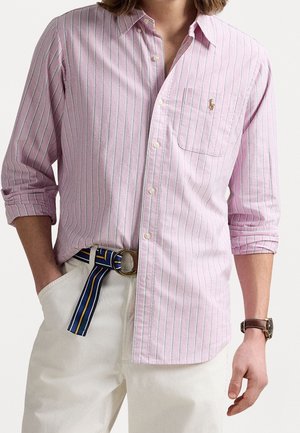 Hombre con una camisa de botones a rayas rosa y blanca con las mangas enrolladas, pantalones blancos, un cinturón azul a rayas y un reloj marrón.