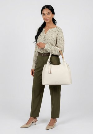 Donna con camicetta beige a fantasia e pantaloni verde oliva che tiene una grande borsa bianca con manici color cuoio su sfondo neutro.