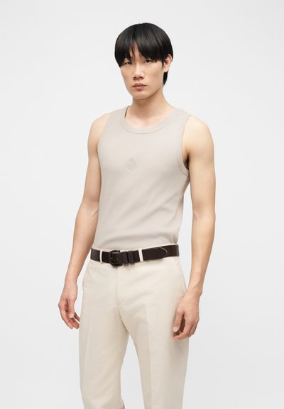 Νεαρός με μαύρα μαλλιά που φορά μπεζ αμάνικη μπλούζα, ανοιχτό μπεζ παντελόνι και σκούρο καφέ ζώνη, στεκόμενος μπροστά σε ένα απλό φόντο.