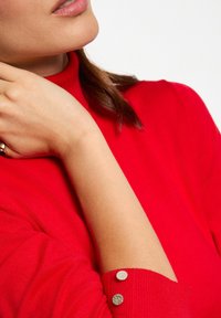 Roter Strickpullover mit hochgeschlossenem Kragen und verzierten Bündchen mit zwei silbernen Knöpfen. Glatte Textur und taillierter Schnitt.