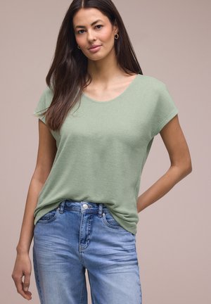 Kvinde med langt mørkt hår iført lysegrøn kortærmet top og blå jeans, stående mod en ensfarvet beige baggrund, den ene hånd på hoften.