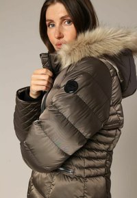 Veste marron rembourrée avec une capuche en fourrure, motif matelassé, poche latérale avec fermeture éclair et patch logo sur la manche. Texture douce.