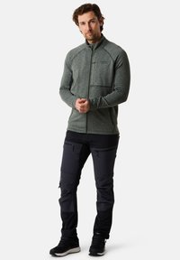 Giacca grigia in pile con zip, tessuto strutturato e tasca frontale, abbinata a pantaloni neri affusolati con tasche con cerniera. Scarpe da ginnastica nere.