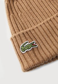Bonnet en tricot marron avec une texture en côtes. Présente un logo de crocodile brodé en vert avec des accents rouges sur un fond blanc.