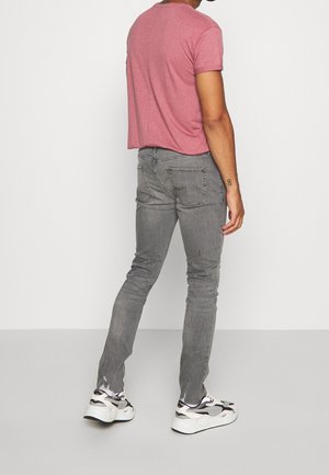 Mann trägt ein ausgebleichtes rotes T-Shirt, graue Slim-Fit-Jeans und weiße klobige Sneaker, steht mit dem Rücken zur Kamera vor einem weißen Hintergrund.