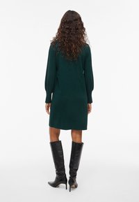 Knee garuma tumši zaļš adīts kleita ar garām pūkainām piedurknēm; pārī ar melnām ceļgalu augstām stiletto zābakām. Gluda tekstūra, vienkāršs dizains.