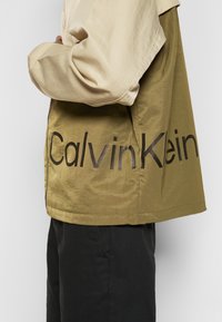 Kurtka Calvin Klein z beżową górną częścią i oliwkowozieloną dolną, gładka w dotyku, z dużym czarnym logo tekstowym po boku.