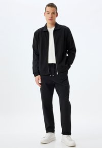 Chaqueta negra con cremallera sobre una camisa blanca, combinada con pantalones negros y zapatillas blancas. La chaqueta tiene un cuello clásico y puños elásticos.