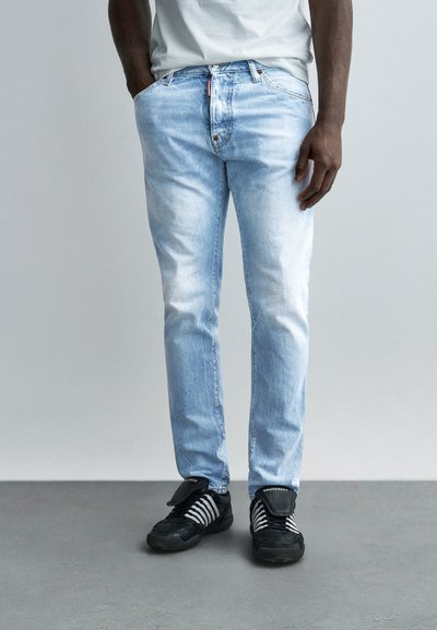 Homme portant un jean bleu clair délavé et des baskets noires, debout avec les mains partiellement dans les poches, devant un fond uni.