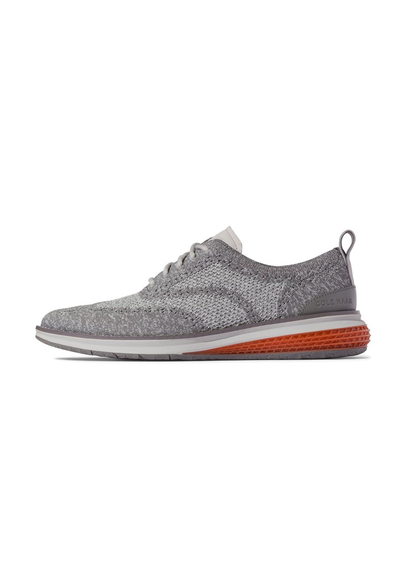 Zapatilla de punto gris con un diseño flexible, que presenta una parte superior texturada, una media suela blanca de perfil bajo y un acento naranja en la suela.