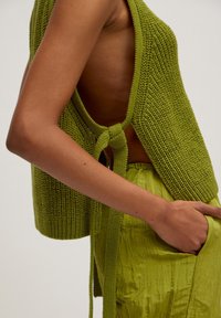 Grünes gestricktes ärmelloses Oberteil mit offenem Rücken, am Bund gebunden. Kombiniert mit zerknitterten grünen Hosen mit seitlichen Taschen und weicher Textur.