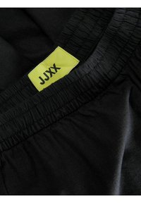 Černé sportovní šortky s elastickým pasem, na kterých se nachází výrazná žlutá nálepka s nápisem "JJXX" a hladkou texturou látky.