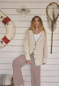 Kremasti kardigan s plavom obrubom i drvenim gumbima, upareni s crveno-bijelim prugastim hlačama, na svijetloj drvenoj pozadini.