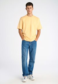 Maglietta di cotone gialla con maniche corte, abbinata a jeans di denim blu. Il modello indossa sneakers bianche con accenti colorati. Sfondo semplice.