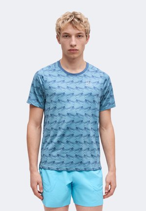 Junger Mann trägt ein blau gemustertes kurzärmeliges Hemd und hellblaue Shorts, steht vor einem schlichten hellgrauen Hintergrund.