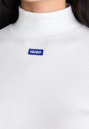 Zbliżenie osoby noszącej biały prążkowany golf z małą niebieską naszywką z napisem HUGO na klatce piersiowej.