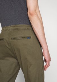 Personne portant un pantalon vert olive avec des poches passepoilées à l'arrière et une chemise gris foncé, main reposant près de la hanche sur un fond uni.