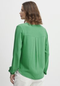 Blusa verde a maniche lunghe con finitura testurizzata, dettaglio di piega sul retro e orlo arrotondato. Indossata con pantaloni chiari.