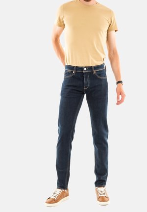 Vaqueros de mezclilla azul oscuro con corte ajustado, con costuras naranjas y cierre de botón, combinados con una camiseta de manga corta beige y zapatillas de color marrón claro.