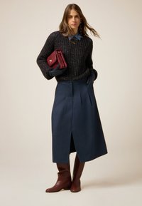 Donna che indossa un maglione nero a trama, una camicia blu con colletto, una gonna midi blu scuro, stivali marroni fino al ginocchio, guanti neri e tiene in mano una pochette rossa.