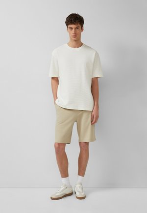 Mann steht an einer grauen Wand, trägt ein weißes kurzärmeliges Hemd, beige strukturierte Shorts, weiße Socken und beige-weiße Turnschuhe.