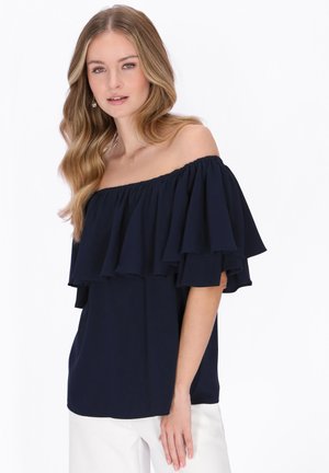 Młoda kobieta z falującymi włosami, ubrana w granatową bluzkę z odkrytymi ramionami i falbankami oraz białe spodnie, pozująca na białym tle.
