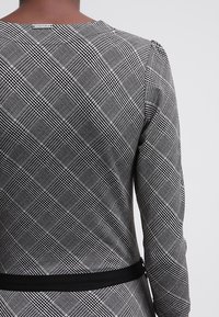 Grå och svart rutigt klänning med långa ärmar, rund halsringning och ett svart bälte, med en strukturerad tyg och synliga sömdetaljer.