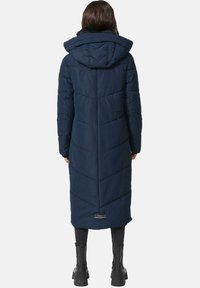 Navyblauwe lange pufferjas met een quiltpatroon, voorzien van een capuchon en contrasterende textuur bij de kraag. Zwarte laarzen zichtbaar aan de onderkant.