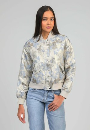 Jeune femme portant une veste bomber bleu clair à fleurs et un jean délavé clair, debout devant un fond gris uni, vue de face.