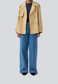 Beige dobbelbrystet trenchcoat med skulderstropper, parret med løse blå denim breddebukser og en mørk overdel. Sorte sko synlige.