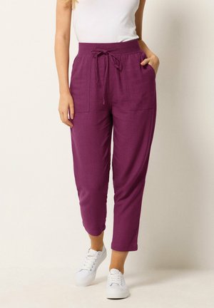 Vrouw draagt paarse broek met hoge taille en trekkoord met voorzakken, een witte mouwloze top en witte sneakers, staande tegen een effen achtergrond.