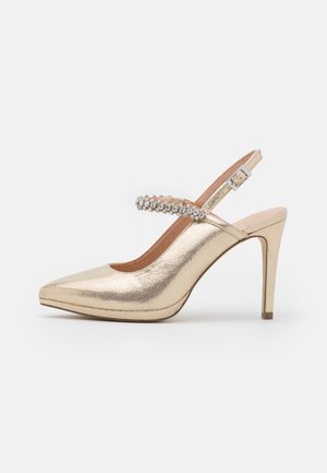 Chaussure à talon haut dorée avec un bout pointu et une bride ornée de strass sur le coup de pied, sur fond blanc.