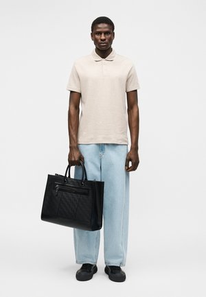 Hombre con camisa polo beige, vaqueros de pierna ancha azul claro, zapatos negros, sosteniendo un gran bolso tote de cuero negro con patrón acolchado y bolsillo con cremallera.