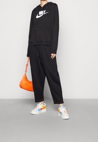 Person som bär en svart Nike-hoodie och byxor, håller en orange handväska och har vita sneakers med röda, gula och blå detaljer.