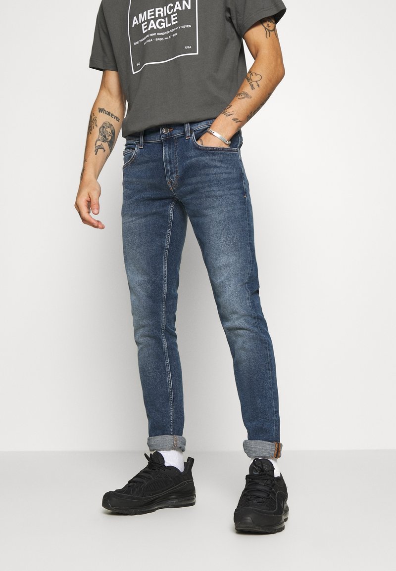 Mörkblå skinny jeans med subtila blekningar, med fem fickor och knappstängning. Kombinerat med en grå grafisk t-shirt och svarta sneakers.
