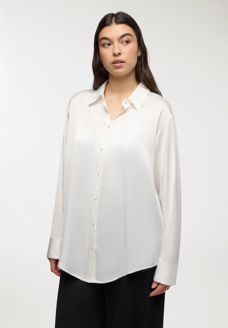 Camicia bianca in satin con maniche lunghe, dotata di colletto, bottoni avorio e orlo curvo. Tessuto liscio e vestibilità ampia.