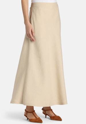 Vrouw die een beige rok tot aan de enkel draagt, gecombineerd met bruine kittenhakken met spitse neus en T-bandje, staand tegen een effen witte achtergrond.